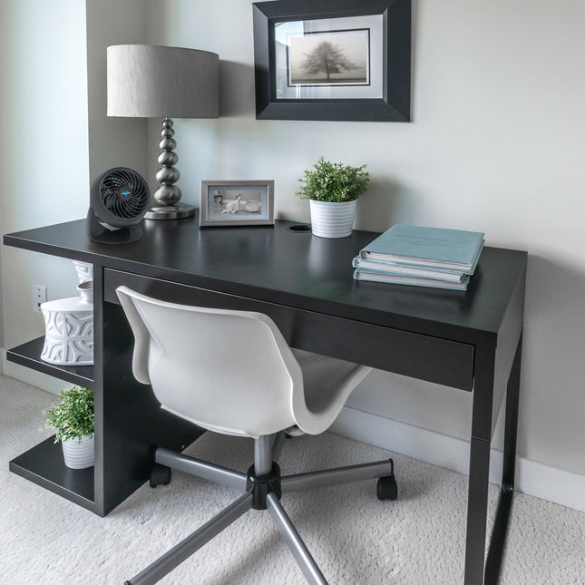 133 air circulator on a black desk in front of a chrome lamp, the desk contains various plants and books and has a gray office chair pushed in