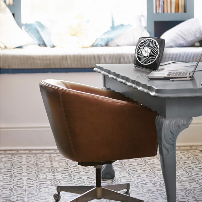 Brown leather office chair in front of a gray desk with a laptop and fan, in a room with a window seat and pillows.