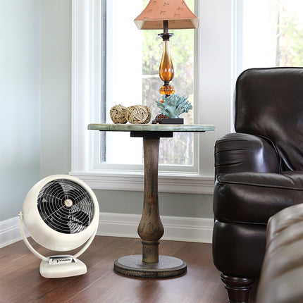 White fan on a wooden floor next to a brown leather chair and a decorative table with a lamp.