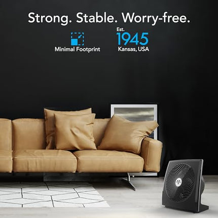 Black fan on a gray floor with a brown couch and black wall in the background.