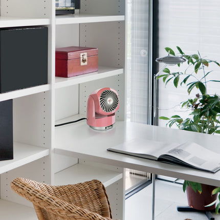 Pink portable fan on a white desk with a book and plant in a room with shelves and a window.