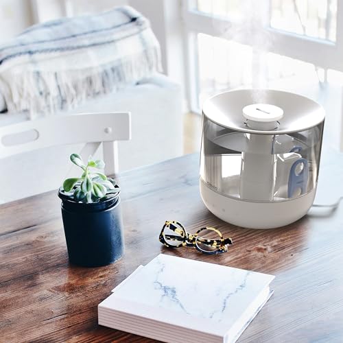 White humidifier on a wooden table with a plant, glasses, and book in a bright room.