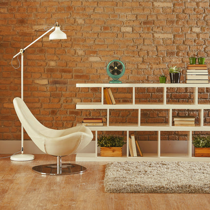 Modern living room with a white chair, floor lamp, and bookshelf against a brick wall.