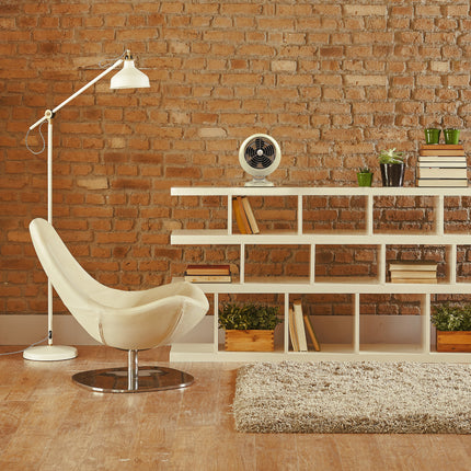 Modern living room with a white chair, floor lamp, and bookshelf against a brick wall.