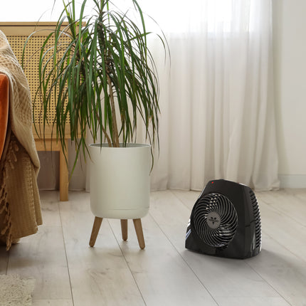 Black space heater on a wooden floor next to a potted plant and chair.