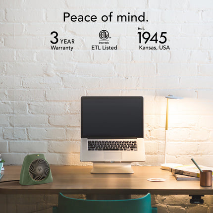 Desk setup with a computer monitor, lamp, and small heater against a white brick wall with brand and product information.