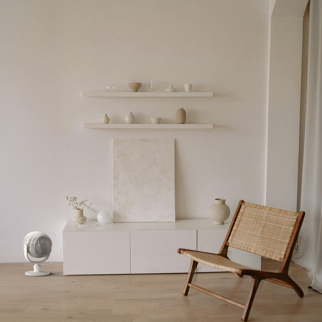Minimalist living room with a wicker chair, white shelves, and a neutral color palette featuring a vornado fan