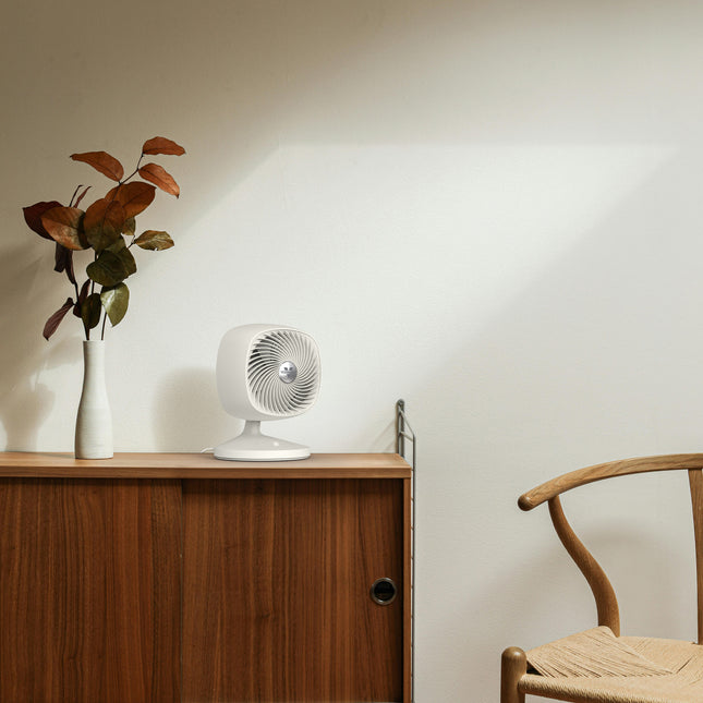 Small white fan on a wooden side table with a plant and chair in the background.