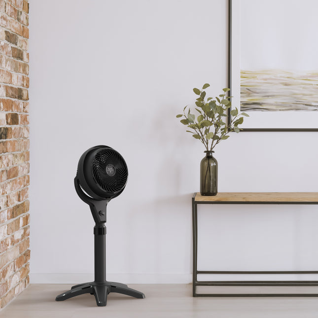 Black oscillating fan on a white floor next to a brick wall and a small table with a vase and plant.