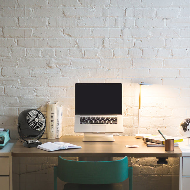 Home office desk with laptop, books, and lamp against a white brick wall.