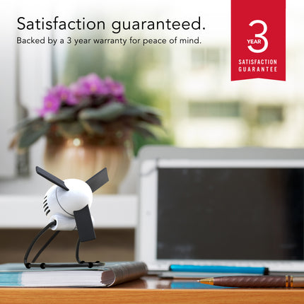 Small white and black fan on a desk with a laptop and plant in the background, featuring a 3-year satisfaction guarantee.