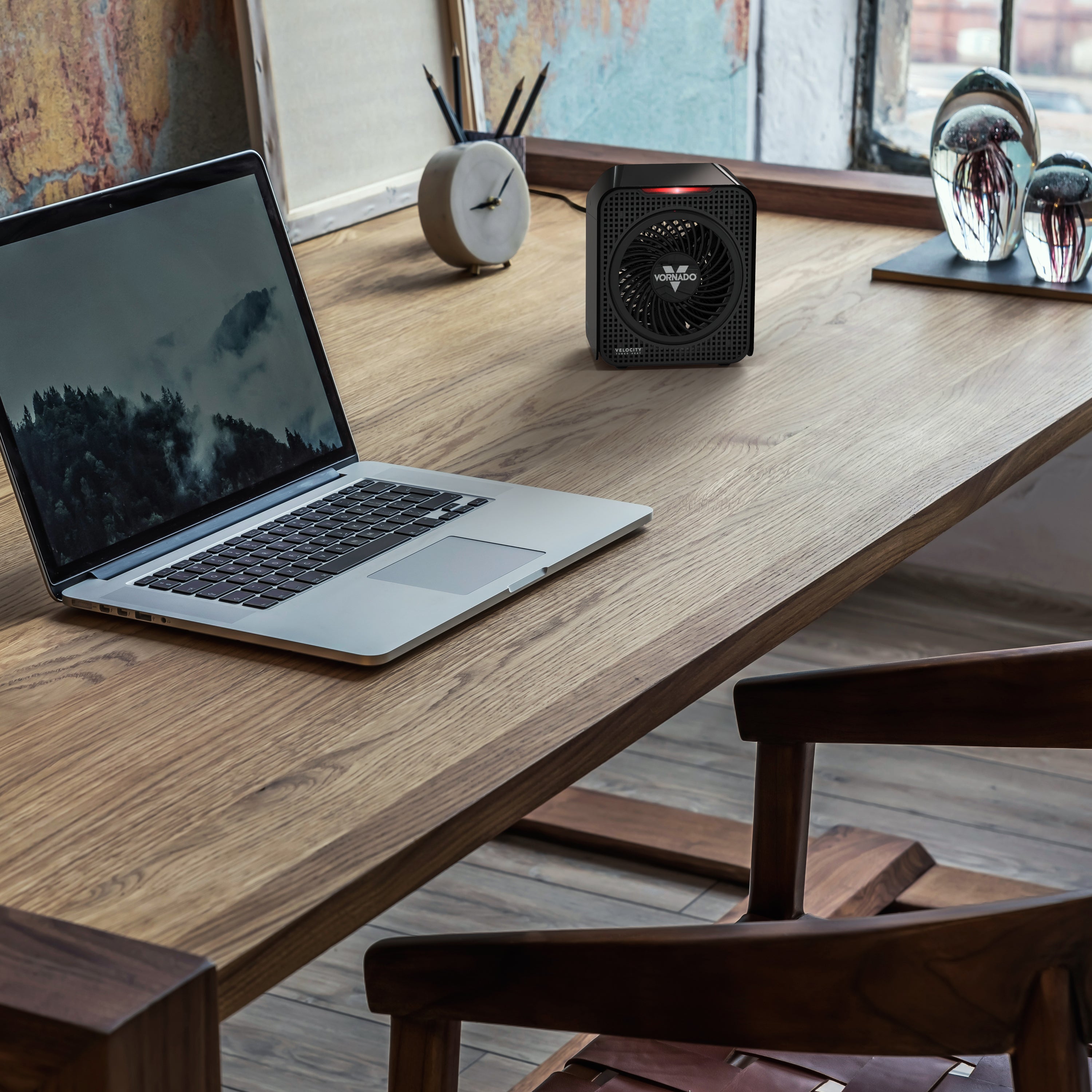 Laptop on a wooden desk with a small black heater next to it, in a room with a window and decor.