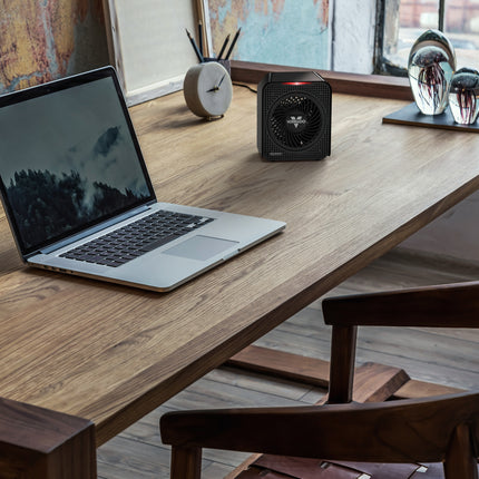 Laptop on a wooden desk with a small black heater next to it, in a room with a window and decor.
