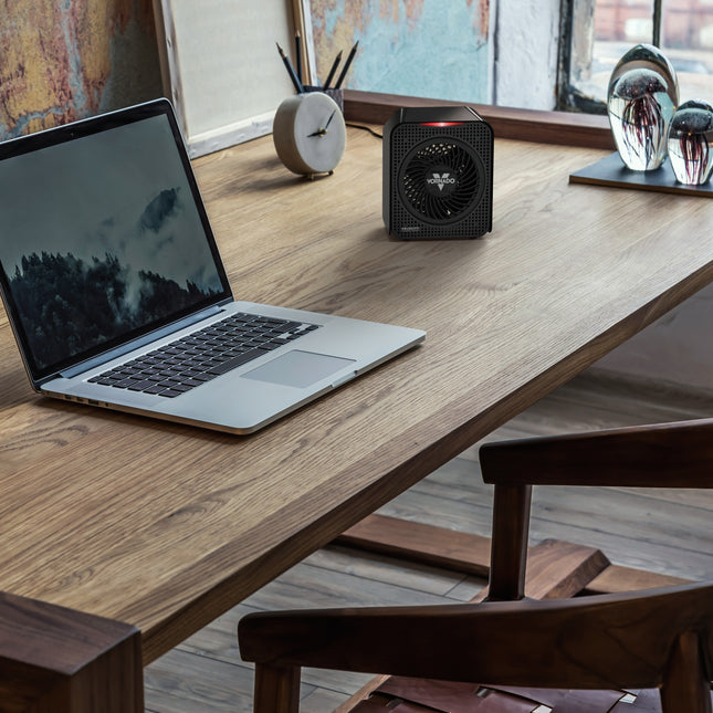 Laptop on a wooden desk with a small black heater next to it, in a room with a window and decor.