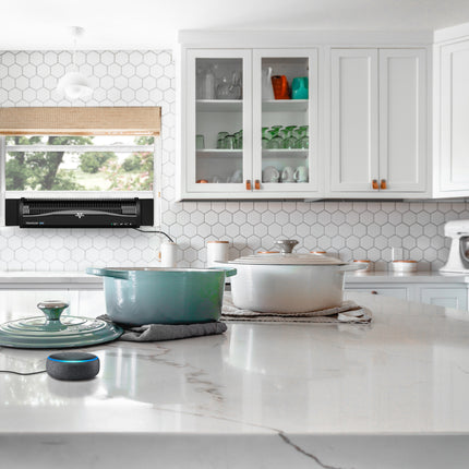 Modern kitchen with white cabinets, marble countertops, and colorful cookware. A black window fan is present in the back window