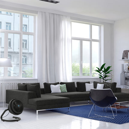Modern living room with a black sectional sofa, blue rug, and a fan on the floor.