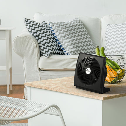 Black fan on a wooden coffee table with a couch and decorative pillows in the background