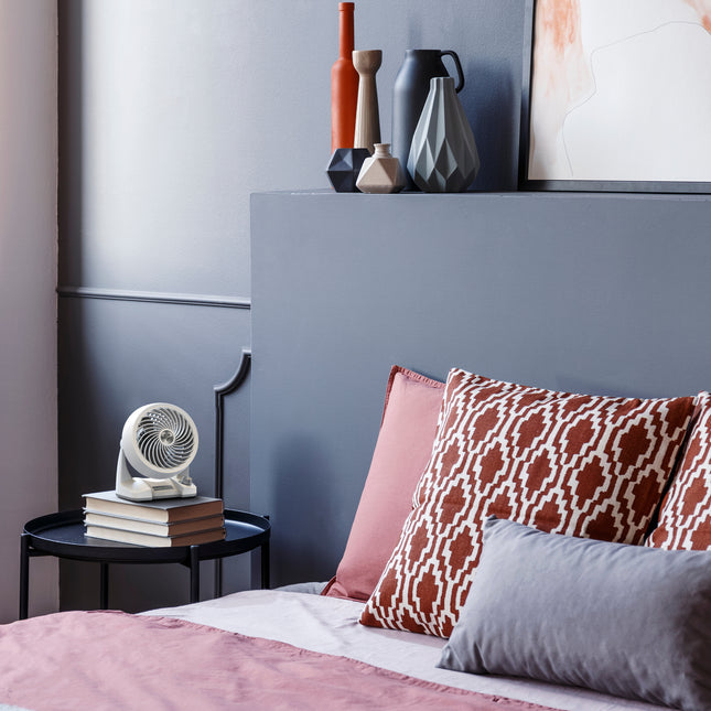 Bedroom with a bed featuring patterned pillows, a nightstand with books and a fan, and decorative vases on a shelf.