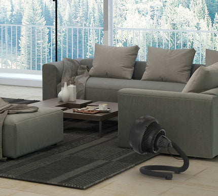 Gray sectional sofa in a living room with a window view of trees, featuring a black portable fan on the floor.