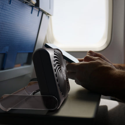 A small black personal fan with a clear plastic stand is placed on an airplane tray table near a window, with a person's hands holding a smartphone nearby.