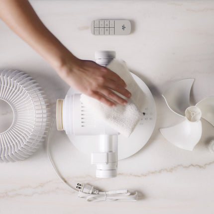 Hand cleaning a white air circulator on a marble surface with a remote control in the background.