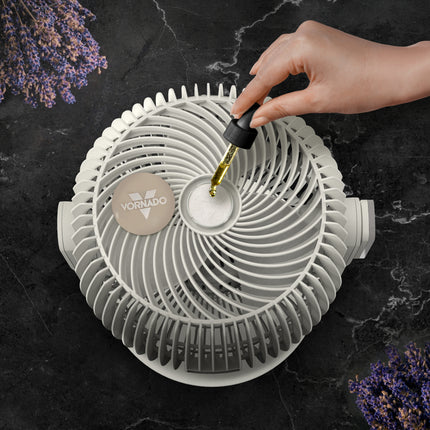 Vornado fan with a hand pouring oil into it on a dark surface with lavender flowers.