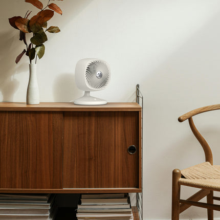 Small white fan on a wooden sideboard with a vase and plant, next to a wooden chair.