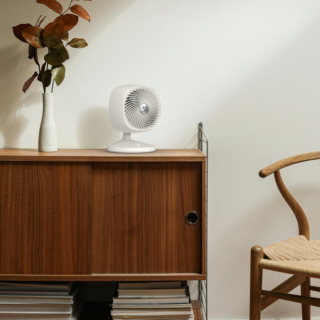 Small white fan on a wooden sideboard with a vase and plant, next to a wooden chair.