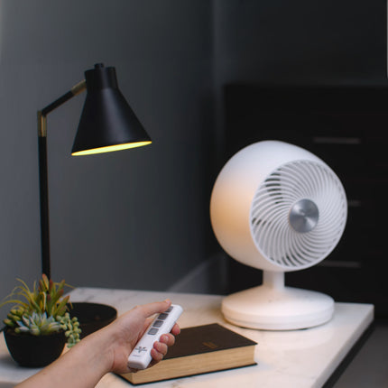 Hand holding a remote control near a white desk fan on a table with a lamp and book in the background.