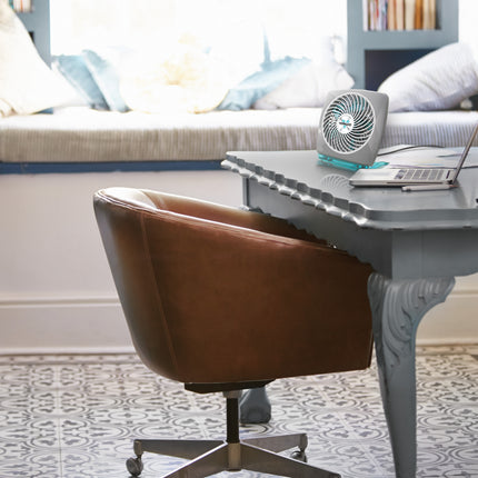 Brown office chair in front of a gray desk with a laptop and fan, in a room with a bed and pillows in the background.