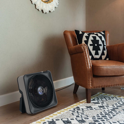 Portable fan on a wooden floor next to a brown leather chair with a patterned cushion.