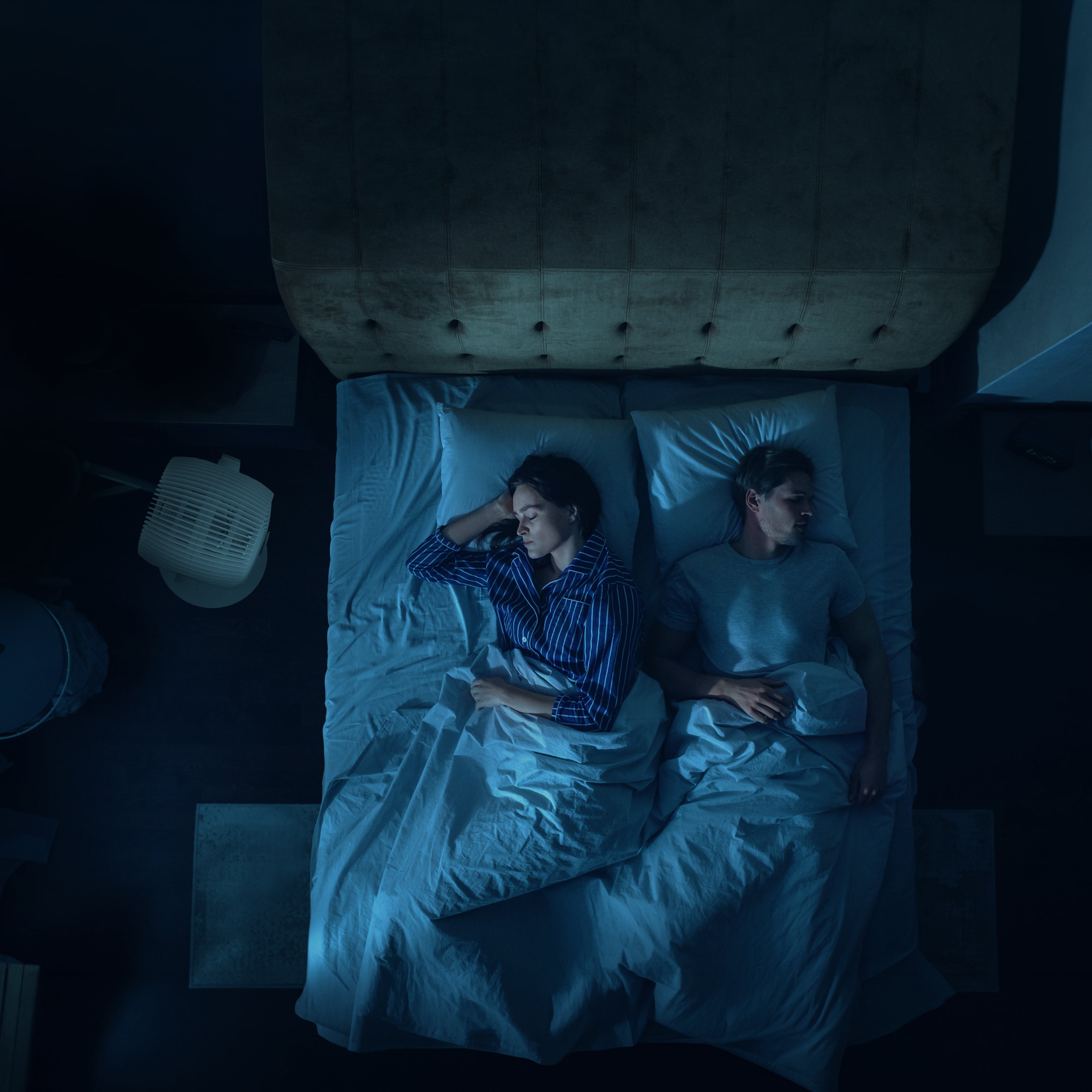 Top-down view of a couple lying in bed in a dimly lit room with an EOS 9S keeping them cool next to the bed.