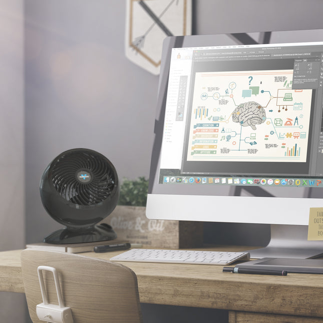 5303 on a wooden desk in front of a plant and nexto to a computer set up with a monitor and keyboard. The walls behind the fan are a light gray color