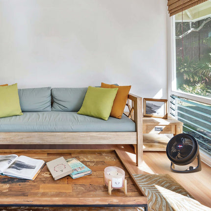 610 in a living room with colorful pillows and a wood framed blue couch. The fan sits in front of a large window and faces a coffee table in front of the couch