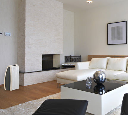 AC550 against a white wall and next to a brick fireplace in a living area. The air purifier faces a white couch and coffee table