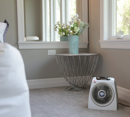 AVH10 on a carpet floor in a bedroom facing a bed with windows behind the heater filling the room with natural lighting