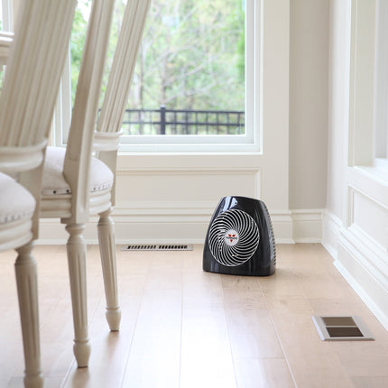 Black space heater on a wooden floor in a room with white chairs and large windows.