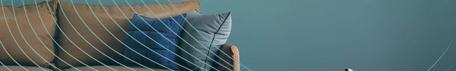 Strata Compact Oscillating Tower Fan on the floor of a living room with a teal wall. There is an orange couch to the left of the fan. Blue lines come out of the fan and go upwards, indicating air circulation.