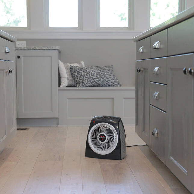Black space heater on a wooden floor in a room with gray cabinets and a window.