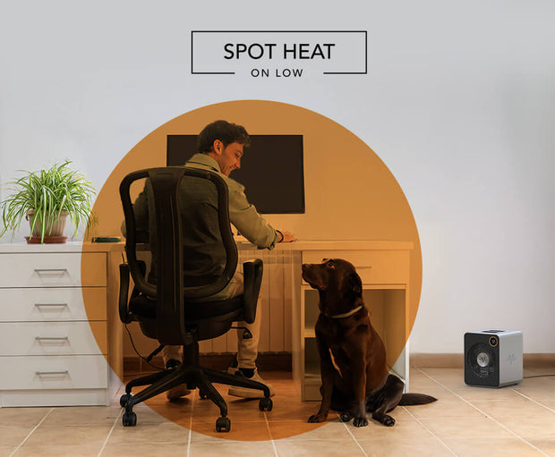 A man sitting at his desk with his dog at his feet. A VMHi600 heater is on the floor nearby. There is an orange circle overlay on him to indicate spot heat on low.