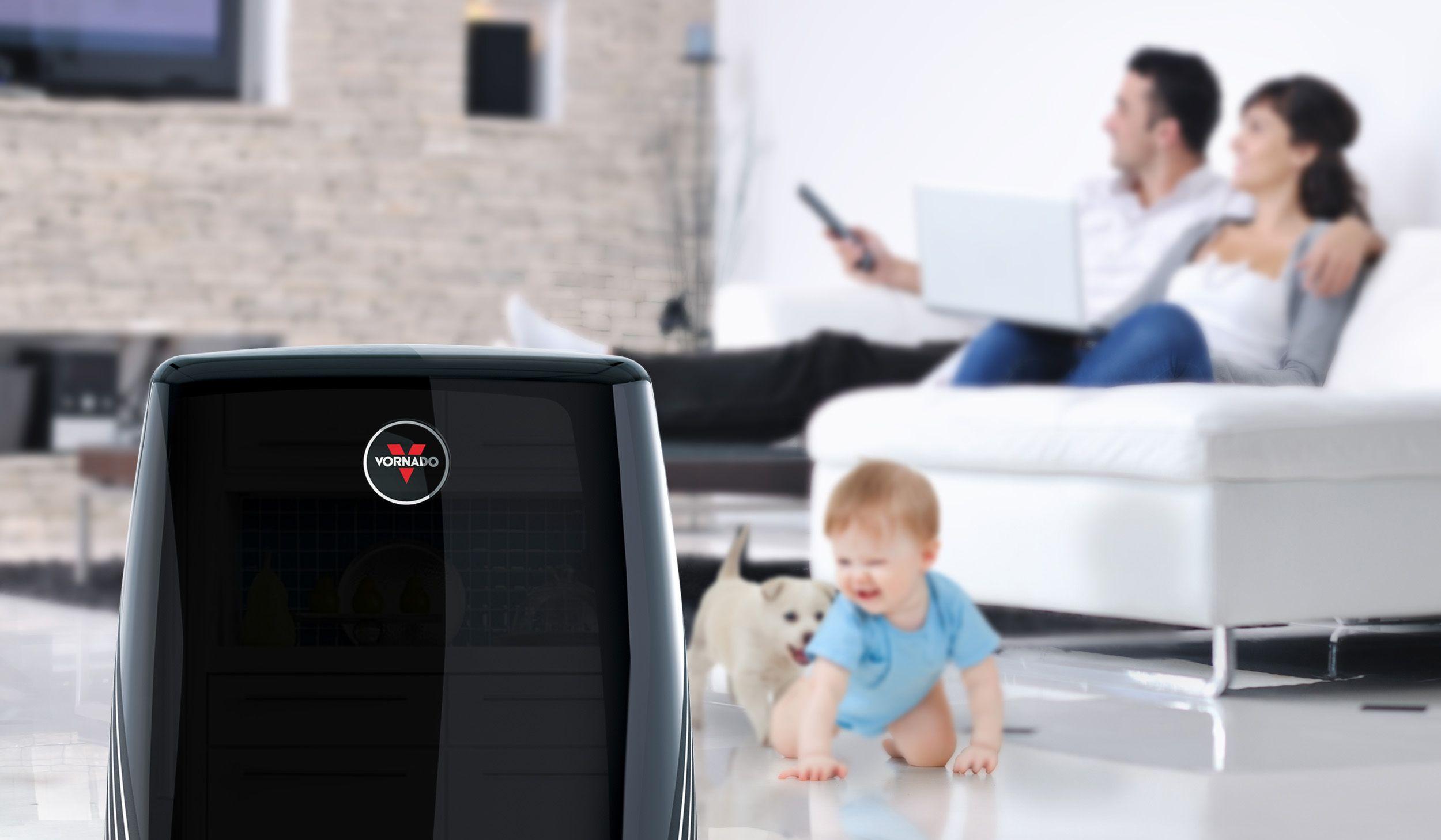 Lifestyle of a black PCO300 air purifier in the foreground. In the background, blurred, is a crawling baby and a puppy. On the couch, the father is changing the channel on the tv and the mother is on her laptop.