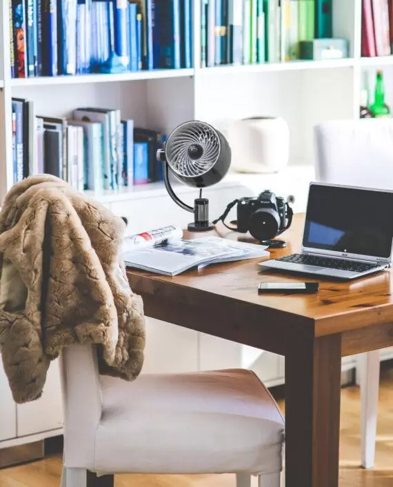 lifestyle of a Black Pivot3 Clip compact Air Circulator on a wood desk. There is a camera, open magazines, and a laptop on the table.