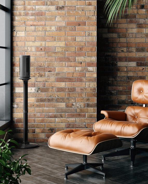 ATOM 1S Compact Oscillating Tower on its stand by a brick wall and leather chair