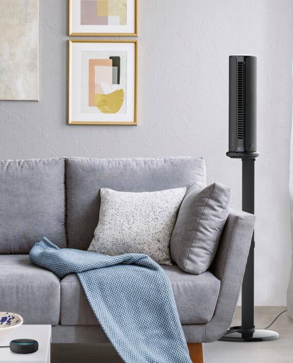 Vornado ATOM 2S AE Compact Oscillating Tower Circulator in Living Room on its stand next to a gray couch. An alexa device in on the table.