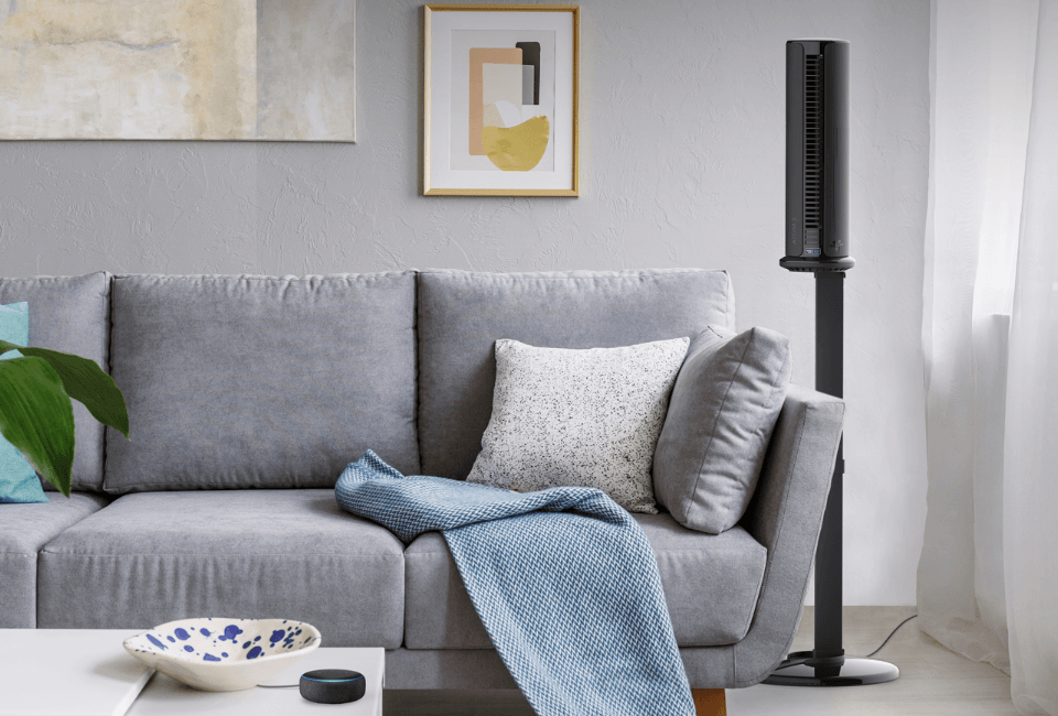 Atom 2S AE Compact Oscillating Tower Circulator with Stand in living room situated next to a gray sofa. An alexa device is on the table by the sofa