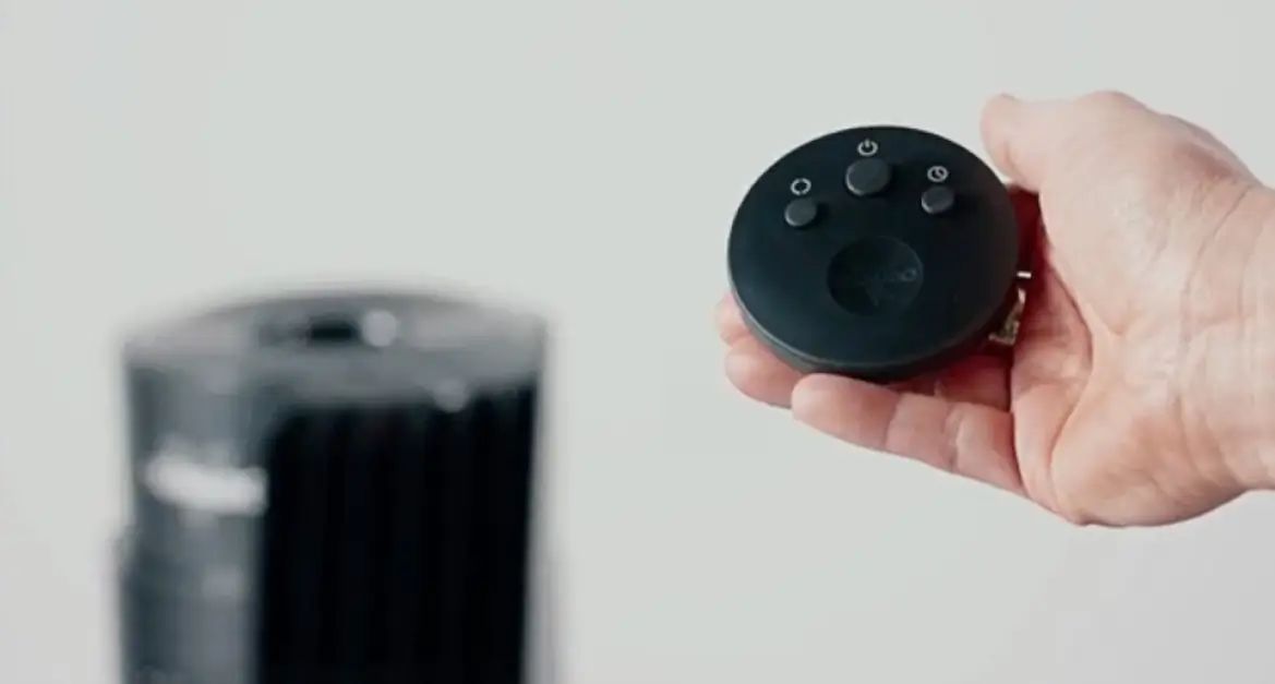 A person holding the OSC54's remote in their hand. The remote is circularly shaped with three gray buttons. The left button is for oscillation, the middle button is the power and fan speed button, and the right button is the timer. In the background is a blurred image of the OSC54 tower fan.