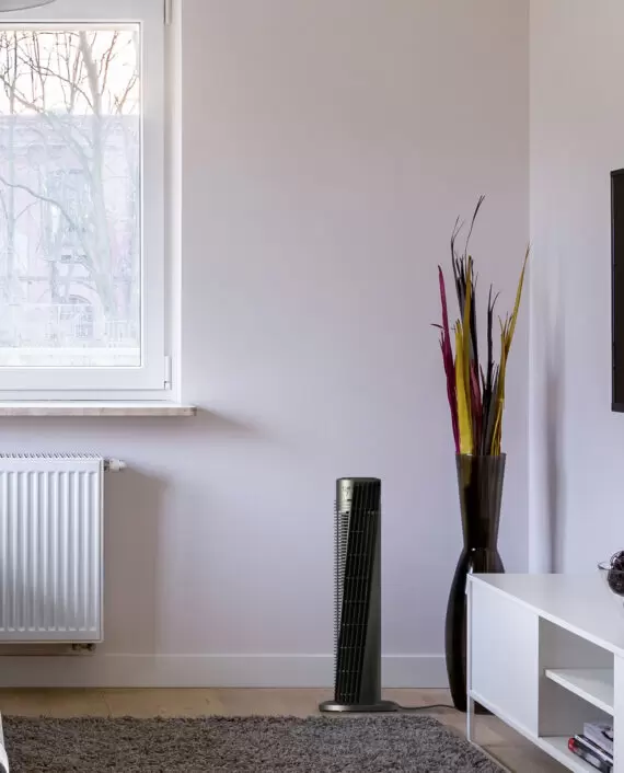 Lifestyle of Vornado OSC54 32" Oscillating Tower Circulator in an apartment next to a window and a large vase with colorful leaves in it.